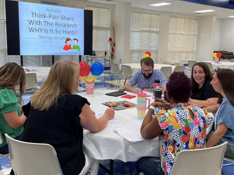 A group of teachers sit around a table during a presentation.